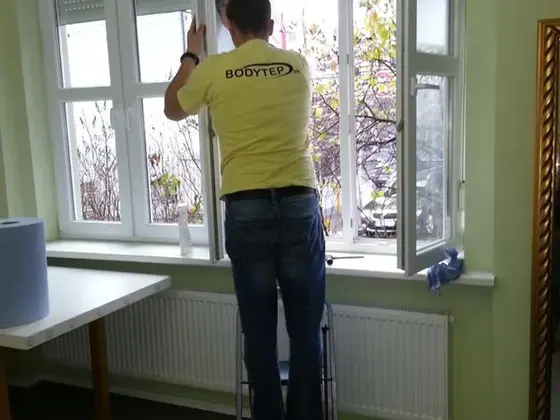 Umývanie okien z malého rebríka: technik v žltom utiera otvorené okno zelenej izby, vedľa rolka utierok