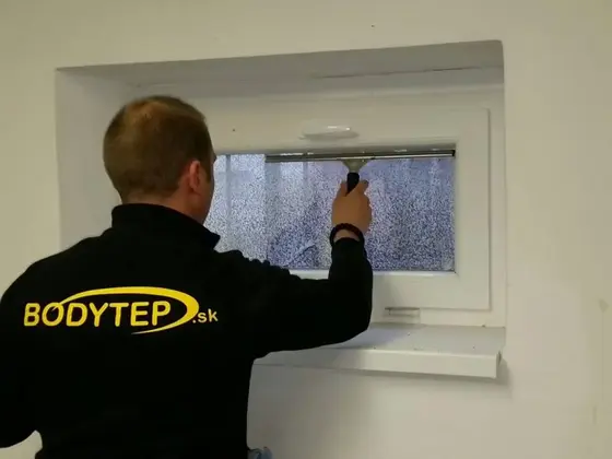 Umývanie sklopného okna v rodinnom dome: technik v čiernej mikine s logom BODYTEP stiera sklo