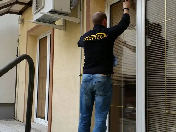 Umývanie okien na terase rodinného domu: technik utiera presklené dvere so žalúziami pod klimatizáciou