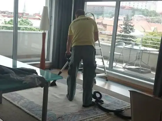 Tepovanie koberca v miestnosti s veľkými oknami: technik pracuje pri sklenenom stolíku, vonku je výhľad na mesto