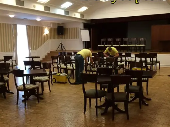 Tepovanie stoličiek v spoločenskej sále: dvaja technici v žltom pracujú medzi stolmi na drevenom parkete