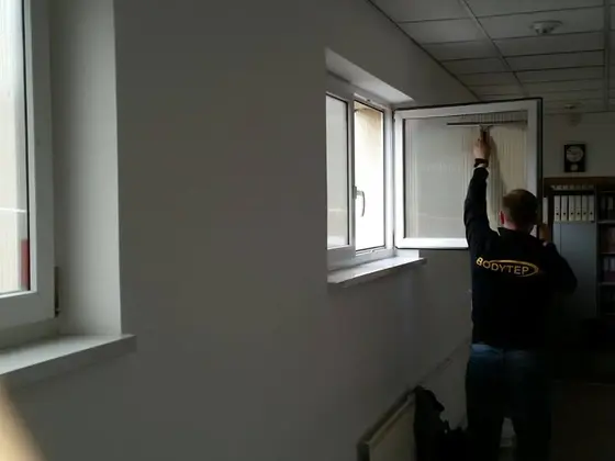 Umývanie okien v Bratislave: technik čistí otvorené krídlo okna so žalúziou, vzadu regály s radmi zakladačov