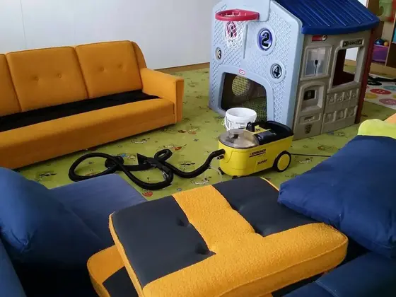 Tepovanie sedačky a kresiel v detskej herni, žltý tepovací stroj stojí na zelenom koberci pri domčeku