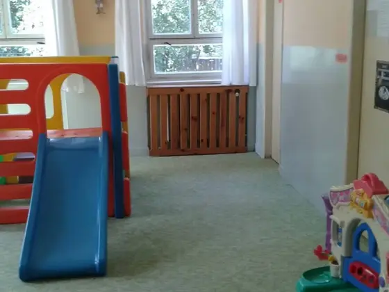 Tepovanie koberca v detskej herni so šmykľavkou a hračkami, zelený koberec a okno s radiátorom