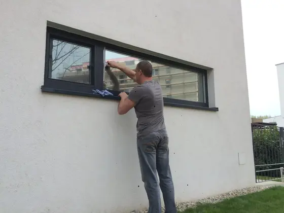 Umývanie okna s tmavým rámom na bielej fasáde, pracovník v sivom tričku utiera sklo, v ňom sa odrážajú budovy