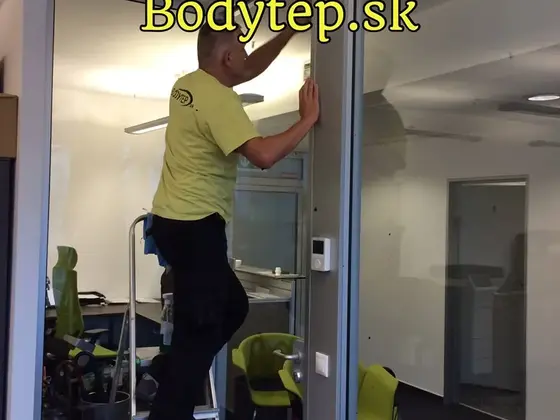 Čistenie presklenia v kancelárii, pracovník na schodíkovom rebríku utiera sklenenú priečku