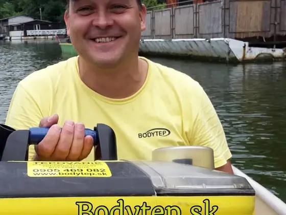 Tepovanie na lodi: usmiaty pracovník v žltom tričku a šiltovke drží tepovací stroj, v pozadí voda