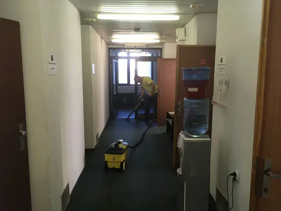 Tepovanie celoplošného koberca na tmavej kancelárskej chodbe, pri stene vodný automat