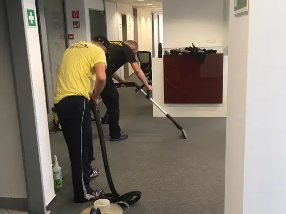 Dvaja pracovníci pri tepovaní sivého koberca v otvorenej kancelárii, na zemi tepovací stroj