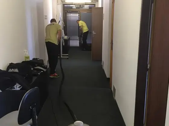 Tepovanie koberca na chodbe kancelárskej budovy: dvaja muži v žltom pri tmavom koberci, vpredu tepovač