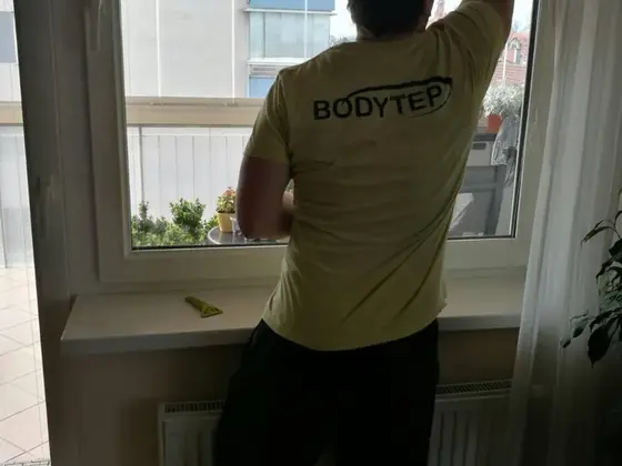 Umývanie okien v byte v Bratislave: muž v žltom tričku stiera horný roh okna, pod oknom radiátor