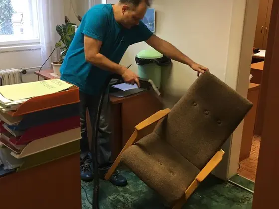 Čistenie kresla v kancelárii: muž v modrom tričku čistí hadicou hnedé čalúnené kreslo s drevenými rámami