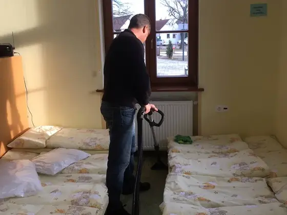 Tepovanie koberca v izbe s lehátkami: muž v tmavom svetri s hadicou, pri okne dva rady lehátok s paplónmi