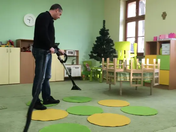 Tepovanie koberca v triede s detským nábytkom: muž s nástavcom medzi zelenými a žltými kruhovými podložkami