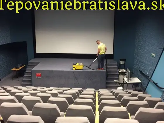 Tepovanie pódia v sále s plátnom: muž s tepovačom na pódiu a pred ním rady sivých sedadiel