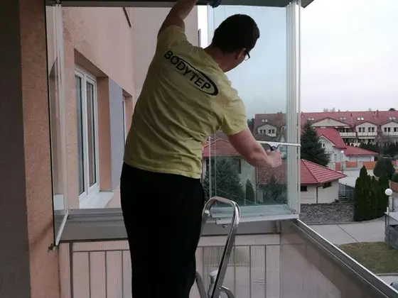 Umývanie okien z rebríka v Bratislave: muž v žltom tričku pri zasklenom balkóne s domami v pozadí