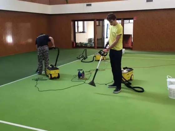 Tepovanie celoplošného koberca v športovej hale so zelenou plochou, dvaja pracovníci s tepovačmi