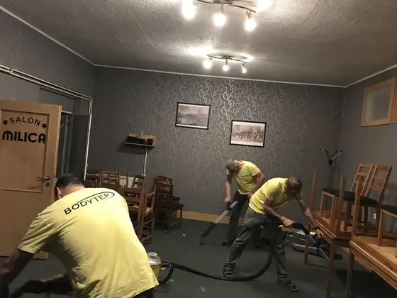 Tepovanie koberca v prevádzke: dvaja technici s hadicou medzi stoličkami, na stenách tmavé tapety