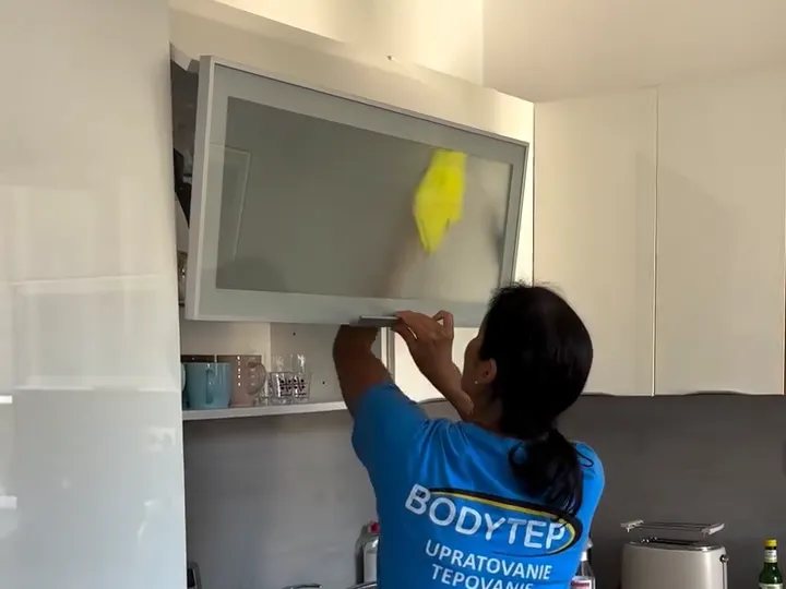 Video: pracovníčka v tričku BODYTEP utiera žltou handrou presklené dvierka hornej kuchynskej skrinky